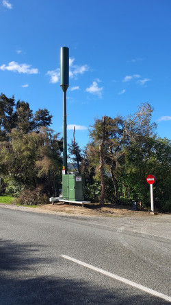 A mobile tower on the side of a road with trees and bush surrounding it.