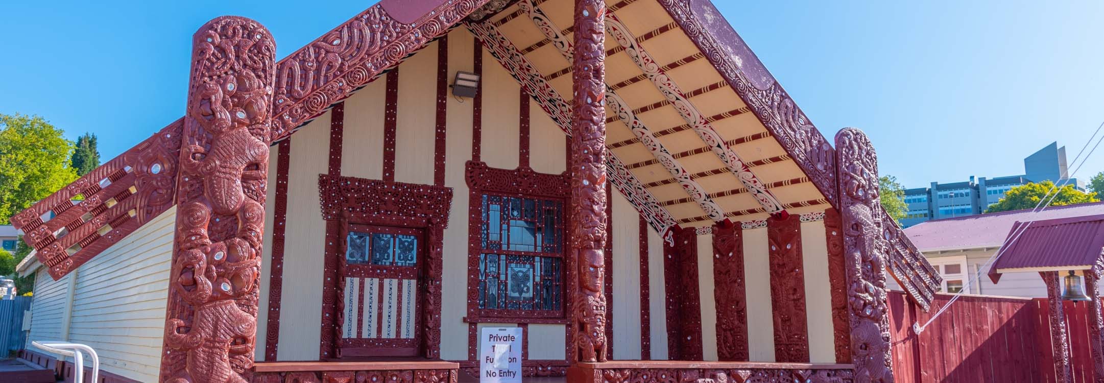 A wharae, part of a marae in front of some trees and blue sky.
