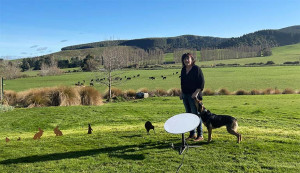 Margie Ruddenklau standing in a grassy paddock with a satellite dish in front of her and a dog by her side.