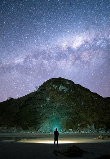A person on a beach silhouetted against a hillside with the milky way visible in the night sky