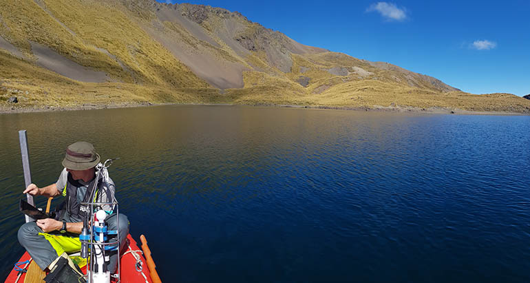 A person sits in a small boat on a still lake. They are using a tablet with other science equipment around them. In the background is a barren mountain scape.