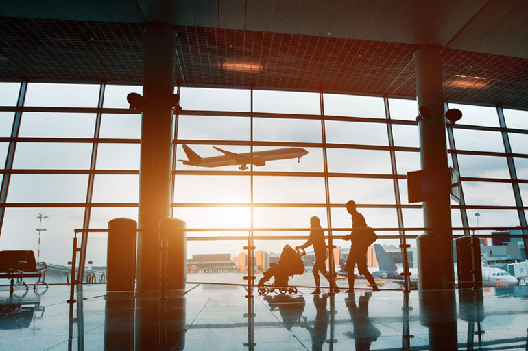 Photograph of a family in silhouette, walking past windows at airport, a plane taking off in background.