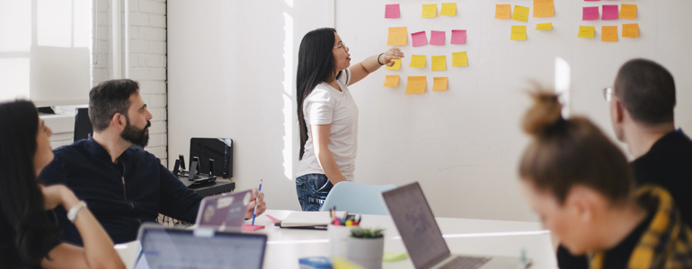 Person explaining sticky notes on a whiteboard to workers sitting at a boardroom table