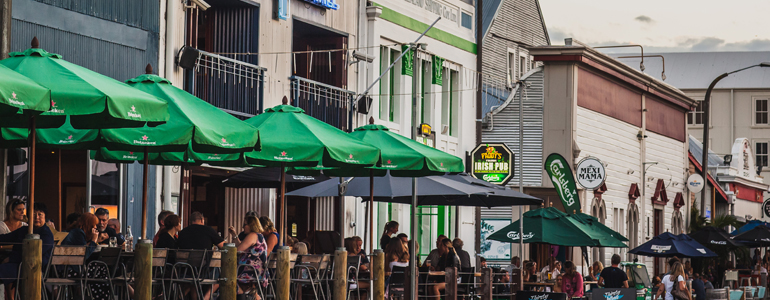 Street front scene with bar patios filled with people drinking under umbrellas.