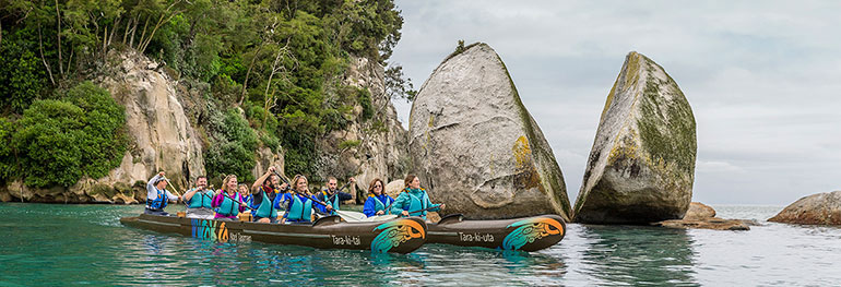 a group of people canoeing at Split Apple Rock in Nelson. Split Apple Rock is a large apple shaped boulder that is split in half, sitting in shallow water on a beautiful beach. 