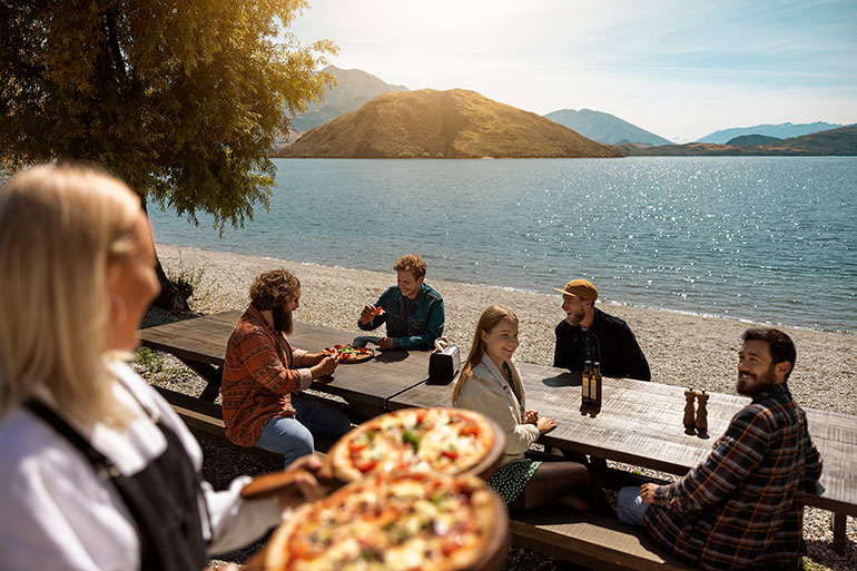Hospitality worker serving a pizza to guests sitting on the shore of a lake.