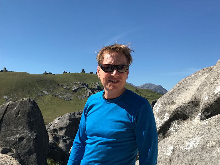 Mark Rocket, wearing sunglasses and bright blue top, stands outside in front of boulders and blue sky.