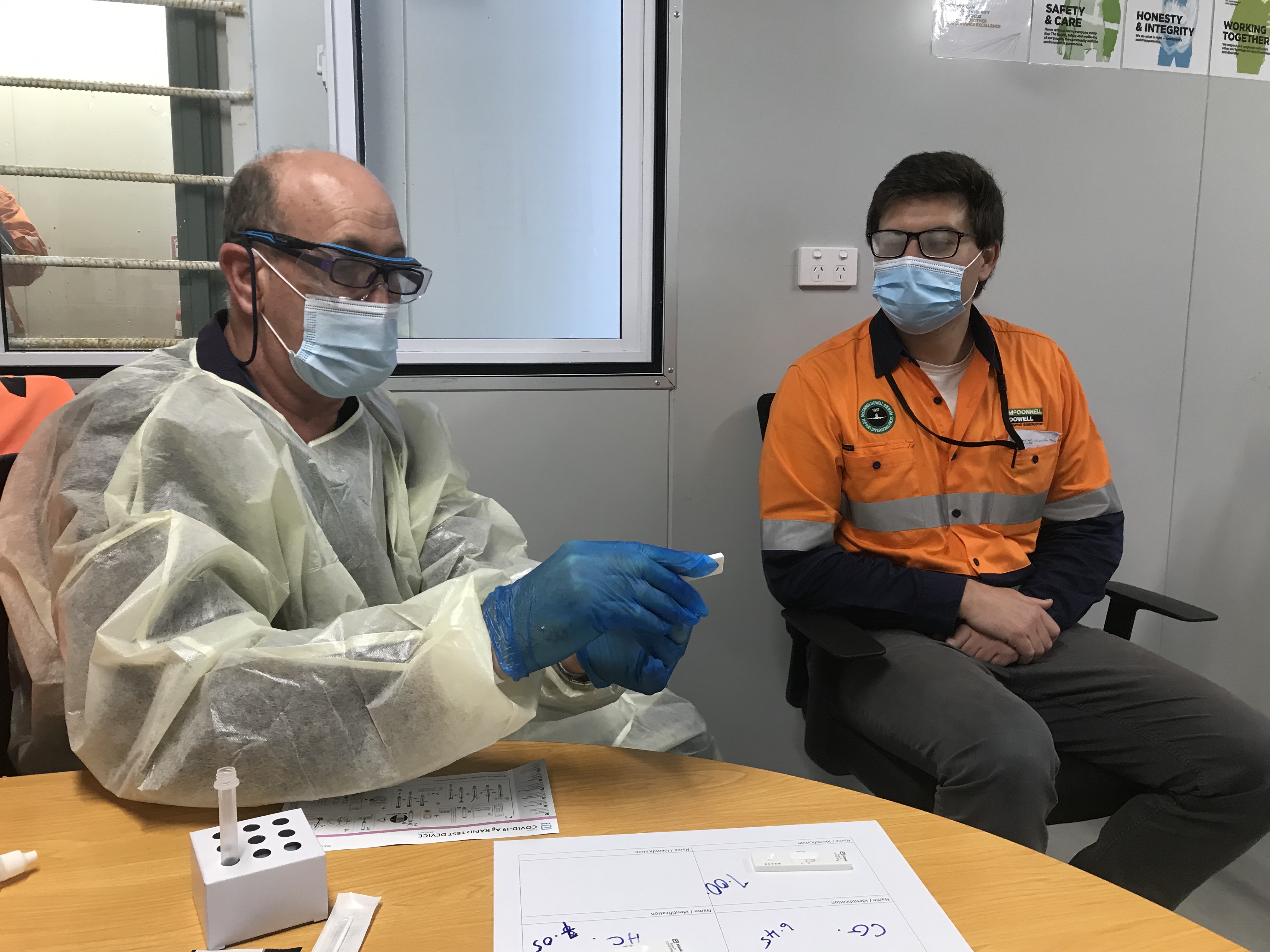 image shows two men in surgical masks sitting in an office ready to give and receive a rapid antigen test.