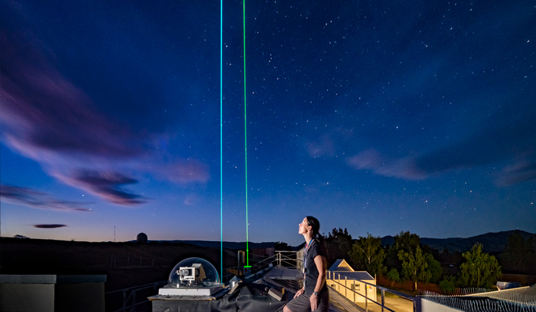 Person looks upwards to a starry night sky. The person is sitting on some sort of platform from which blue and green light is beaming.